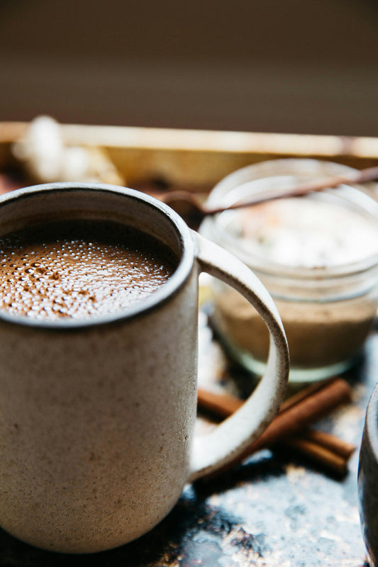 A mug of rich hot chocolate topped with whipped cream and a cinnamon stick, cozy blanket in soft-focus background, warm indoor light.