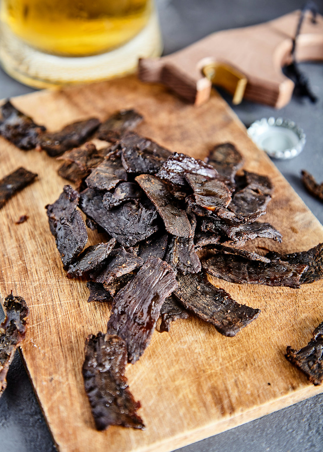 A rustic wooden board with assorted dried meat snack strips, outdoor or hiking lifestyle setting, natural daylight.
